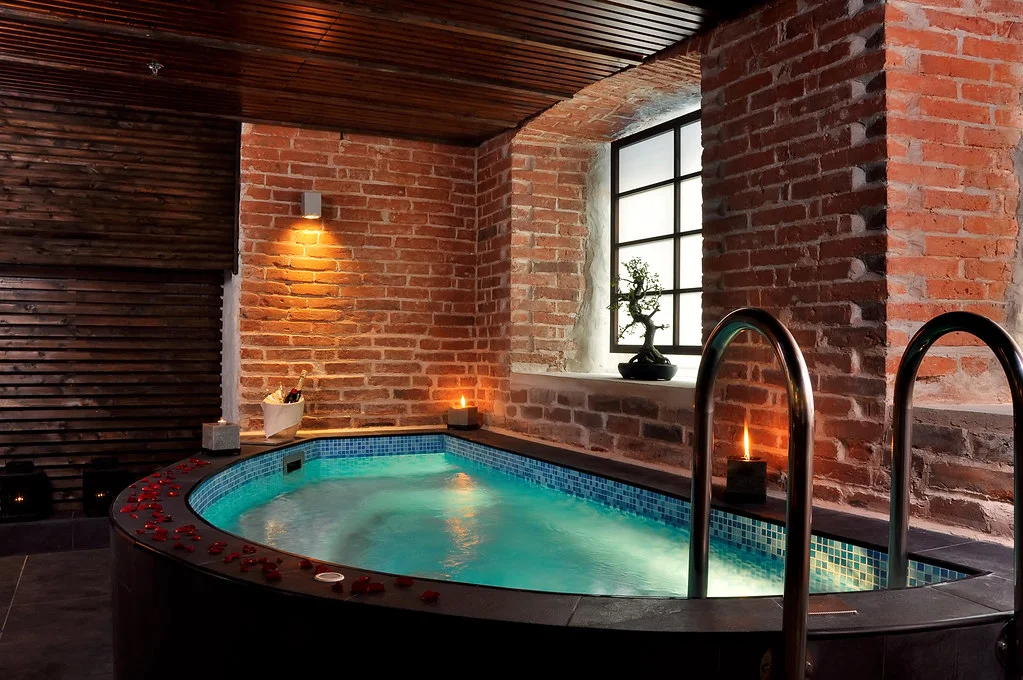 Bassin de spa privatif chauffé entouré de briques, de bougies et de pétales de rose dans une ambiance feutrée.